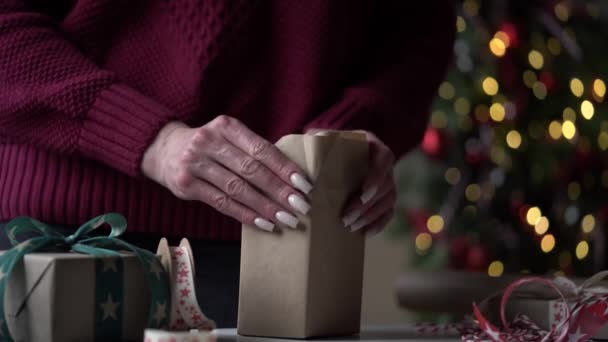 バーガンディセーターのクローズアップ女性は 美しい紙やリボンで贈り物と箱の多くをパックし クリスマスツリーの背景に弓します クリスマス ギフトラッピングの準備 — ストック動画