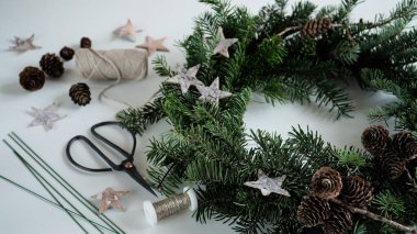 Noel çelengi kapısı, yıldızlarla süslenmiş doğal Noel ağacı dalları, koniler, makas ve kurdeleler beyaz masa, üst görünüm, kopyalama alanı. Ev için Noel çelengi yapma kavramı..