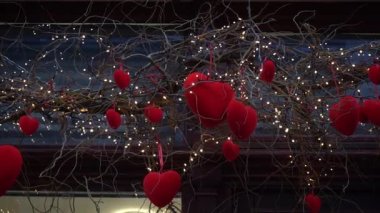 Close-up of red velvet hearts on willow branches among garland of lanterns,festive decor for Valentine's Day.Beautifully decorated shop window in the city.Cityscape, hearts in snow. Slow motion 4K