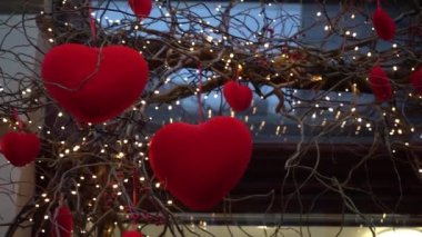 Close-up of red velvet hearts on willow branches among garland of lanterns,festive decor for Valentine's Day.Beautifully decorated shop window in the city.Cityscape, hearts in snow. Slow motion 4K