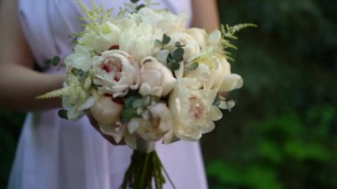 Beyaz elbiseli gelin elinde güzel bir buket beyaz şakayık, astilbe, okaliptüs yeşil ağaçların arka planında. Düğün çiçekçisi, gelin buketi, narin çiçek..