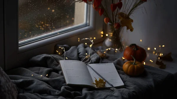 Beautiful atmospheric photograph of autumn mood.Vase with branches of orange physalis, pumpkins,light garland,blanket and book on windowsill near window wet from rain.Autumn, fall, hygge home decor