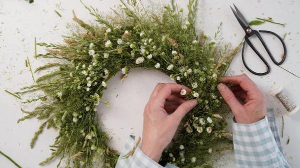 Floristic master class on weaving flower wreath for feast of Ivan Kupala.Woman weaves wreath of fresh field herbs and flowers,pagan symbol,solstice day.Rustic style.Wreath of daisies and herbs,summer