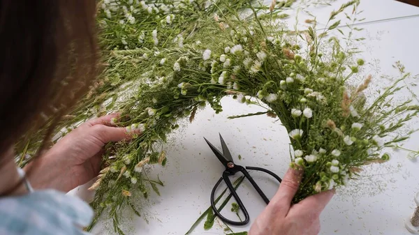 Floristic master class on weaving flower wreath for feast of Ivan Kupala.Woman weaves wreath of fresh field herbs and flowers,pagan symbol,solstice day.Rustic style.Wreath of daisies and herbs,summer