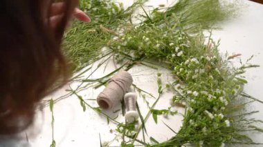 Ivan Kupala şöleni için çiçek dokuma konusunda usta bir sınıf. Kadın taze otlar ve çiçeklerden çelenk dokuyor, pagan sembolü, gündönümü günü. Kırsal stil. Papatya ve bitki çelengi, yaz tatili.