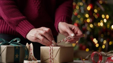Burgonya rengi süveterli bir kadın, içinde hediyeler olan bir sürü kutuyu güzel kağıt ve kurdelelerle paketliyor, bir Noel ağacının arka planında fiyonklar. Noel, yeni yıl tatili, hediye paketi..