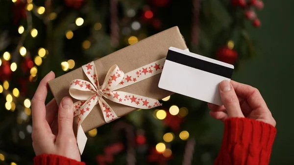 Hands of a woman in a red sweater holding a credit card and a gift box on the background of a Christmas tree close-up. Christmas and New Year shopping online, credit card payment. Buying gifts online