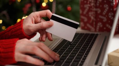 Elinde kredi kartı olan genç bir kadın dizüstü bilgisayarla Noel dekoru ve hediyelerinin arka planına karşı alışveriş yapıyor. Yakın plan. Noel ve Yeni Yıl çevrimiçi alışveriş, kredi kartı ödemeleri.