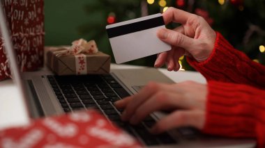 Elinde kredi kartı olan genç bir kadın dizüstü bilgisayarla Noel dekoru ve hediyelerinin arka planına karşı alışveriş yapıyor. Yakın plan. Noel ve Yeni Yıl çevrimiçi alışveriş, kredi kartı ödemeleri.