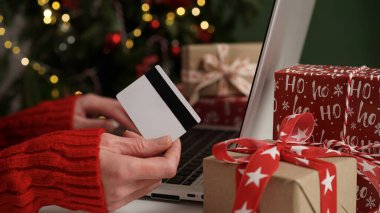Elinde kredi kartı olan genç bir kadın dizüstü bilgisayarla Noel dekoru ve hediyelerinin arka planına karşı alışveriş yapıyor. Yakın plan. Noel ve Yeni Yıl çevrimiçi alışveriş, kredi kartı ödemeleri.