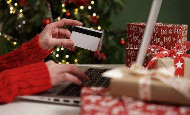 Elinde kredi kartı olan genç bir kadın dizüstü bilgisayarla Noel dekoru ve hediyelerinin arka planına karşı alışveriş yapıyor. Yakın plan. Noel ve Yeni Yıl çevrimiçi alışveriş, kredi kartı ödemeleri.