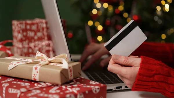 Young woman holding a credit card and makes purchases on a laptop against the background of Christmas decor and gifts, close-up. Christmas and New Years online shopping, credit card payments.