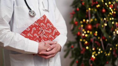 Noel ve Yeni Yıl için tıbbi afiş konsepti. Beyaz önlüklü kadın doktor, arka planda güzel dekore edilmiş Noel ağaçlarına karşı kırmızı kağıda sarılı steteskop ve hediye kutusu tutuyor. Boşluğu kopyala