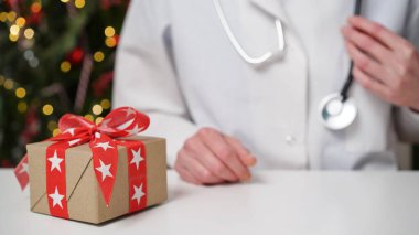Noel ve Yeni Yıl için tıbbi afiş konsepti. Beyaz ceketli kadın doktor masasında oturuyor. Masasında kırmızı fiyonklu hediye kutusu var.