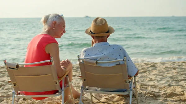 夏休みにはビーチで太陽と海を眺めながら椅子に座っているお年寄りのカップルが笑顔で休暇を楽しんでいます — ストック写真