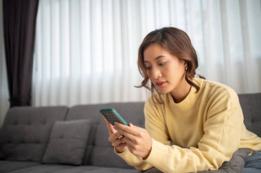 Asyalı kadın akıllı telefon kullanıyor, interneti tarıyor ve evdeki kanepede alışveriş yapıyor.