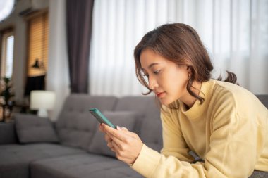 Mutlu genç Asyalı kadın kanepede rahat rahat oturup akıllı telefonuna mesaj atıyor, gülümseyen kız akıllı telefon konuşmaları yapıyor, cihazda kablosuz internet kullanıyor, evden online alışveriş yapıyor.