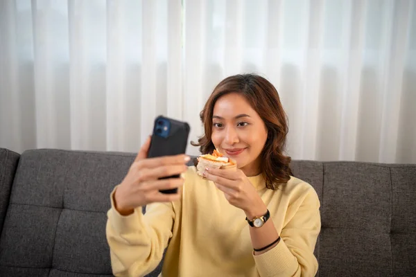 Asian woman using phone video call with friend while eating sandwich at home