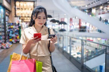 Gündelik giyinen genç bir kadın alışveriş merkezinde kredi kartıyla online alışveriş torbaları taşıyor.