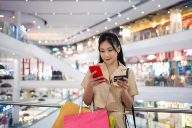 Gündelik giyinen genç bir kadın alışveriş merkezinde kredi kartıyla online alışveriş torbaları taşıyor.