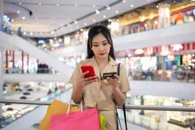 Gündelik giyinen genç bir kadın alışveriş merkezinde kredi kartıyla online alışveriş torbaları taşıyor.