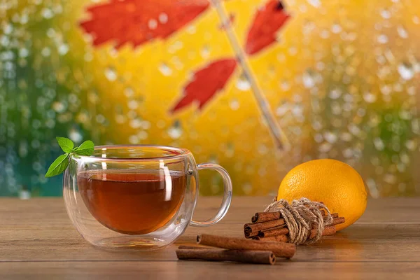 Hot tea in a transparent double glass cup on the table with mint leaves, cinnamon, and lemon with a warm autumn background.