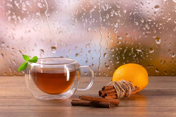 Hot tea in a transparent double glass cup on the table with mint leaves, cinnamon, and lemon near the rainy autumn window background.