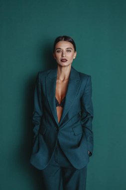 Zarif yeşil takım elbiseli, ceketli ve yeşil duvarda duran pantolonu olan şık, kendine güvenen bir kadın. Stüdyo moda portresi