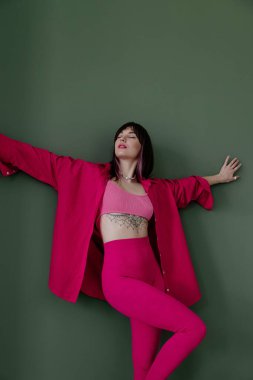 Yeşil duvarın yanında poz veren, pembe dövmeli, modaya uygun beyaz bir kadın.