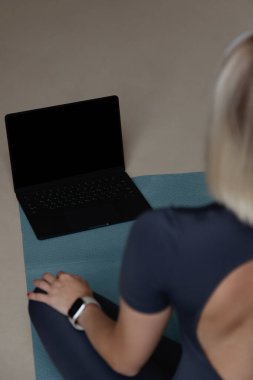 Genç sportif spor spor spor eğitmeni kadın yoga egzersizleri yaparken siyah ekranlı dizüstü bilgisayar kullanarak evin içinde mavi paspas döşemesinde oturuyor.