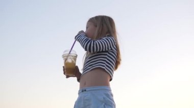 Moda videosu. Yelekli küçük bir kız yazın gökyüzüne karşı portakal suyu içer. Gün batımı gökyüzü. Yaz Tatili 'nde gökyüzünde kız portresi. Yüksek kaliteli FullHD görüntüler