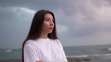 Okyanusun üzerinden gün batımına bakan koyu siyah saçlı, Ciddi Genç Kadın portresi. Bulutlu yağmurlu bir gün, yavaş çekim. Güzel bir kadın uzaklara bakar.