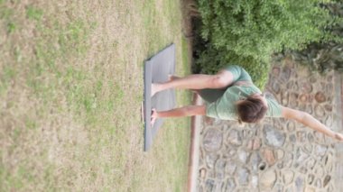 Bir kadın parkta çimlerin üzerinde yoga yapıyor, yazın sokakta esneme antrenmanı yapıyor. Dikey Video
