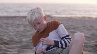 Close-up, a handsome blond boy sitting on the beach by the sea in autumn, Turkey, Alanya.