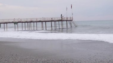 A small storm at sea at the pier, in the background a fisherman with a fishing rod catches fish on a wooden pier going into the sea.