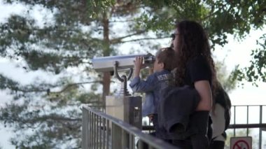 View from the back, tourists mother and son watching binoculars on a walk in the park.