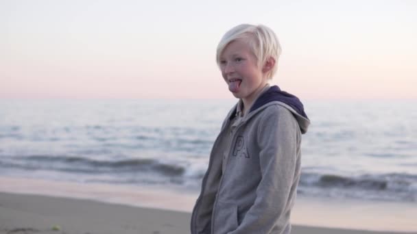 白髪の少年は日没時に海のそばのビーチで舌を突き出す — ストック動画