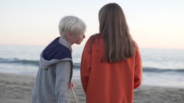 長い黒い髪をした金髪の男の子と女の子がカメラに背を向けて立ち 海を見ている コンセプト 日付の愛のティーンエイジャーの子供たち — ストック動画