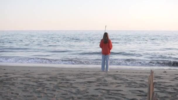 ジーンズとスウェットシャツを着た美しいブルネットの女の子は捨てられたビーチで海を見渡す — ストック動画