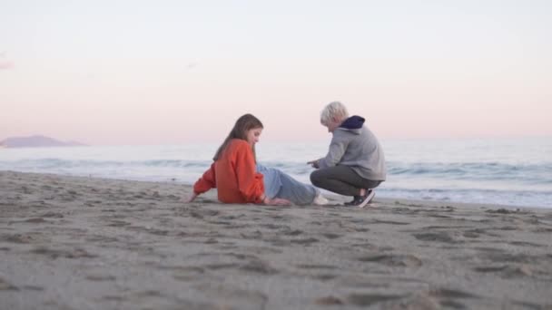 小さな男の子と11歳の女の子は海のそばのビーチに座って話をする — ストック動画