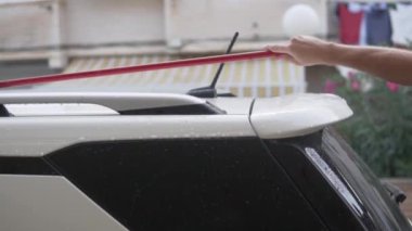 Kirli beyaz bir arabanın çatısını evin yanında paspasla yıkayan bir adam..