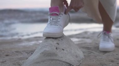 Yakın plan, deniz kenarındaki bir taşın üzerinde ayakkabısına ayakkabı bağı bağlayan bir kadın..