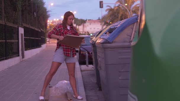 一个女人晚上把垃圾扔进街上的垃圾桶里 一个女孩把空的纸板箱从家具下面扔出去 — 图库视频影像