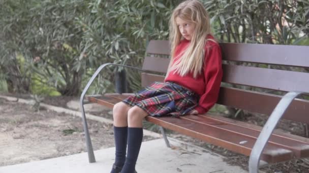 穿着校服的美丽而悲伤的金发姑娘坐在公园的长椅上 — 图库视频影像