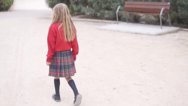 从后面看去 一个穿着格子裙的一年级学生和一件红色毛衣穿过公园步行回家 西班牙 阿利坎特 — 图库视频影像