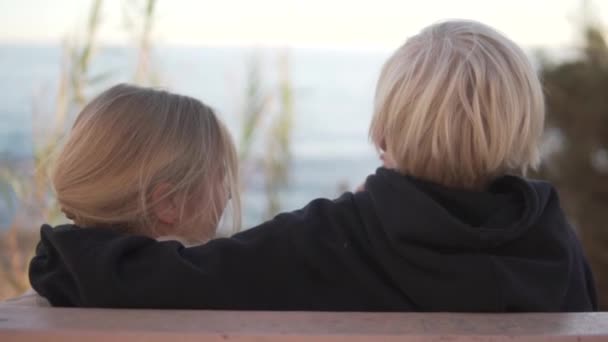 一个男孩和一个女孩坐在海边的长椅上 一个金发女孩抱着一个金发男孩 — 图库视频影像