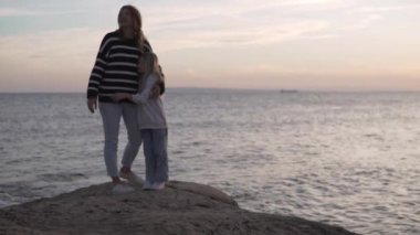 Anne ve küçük kızı, akşam vakti deniz kenarında bir kucaklaşmada dururlar..