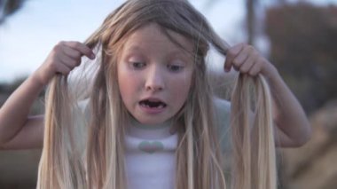 Komik küçük kız gördüğü şeye şaşırmış bir şekilde saçını çekiyor. Uzun sarı saçlı küçük kız neşeli bir şekilde saçlarını elinde tutuyor ve mutlu bir jest yapıyor. Dışarıda çok eğleniyor.