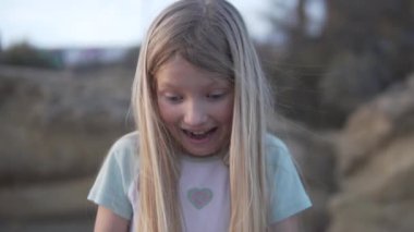Uzun, katmanlı sarı saçlı mutlu küçük bir kız aşağıya bakarken gülümsüyor. Sörfçü saçı arka planda çim ve ahşap manzarasına ekleniyor.