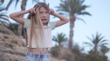 Palmiye ağaçları ve gökyüzü üzerinde dans eden uzun sarı saçlı neşeli küçük kız, İspanya, Alicante.